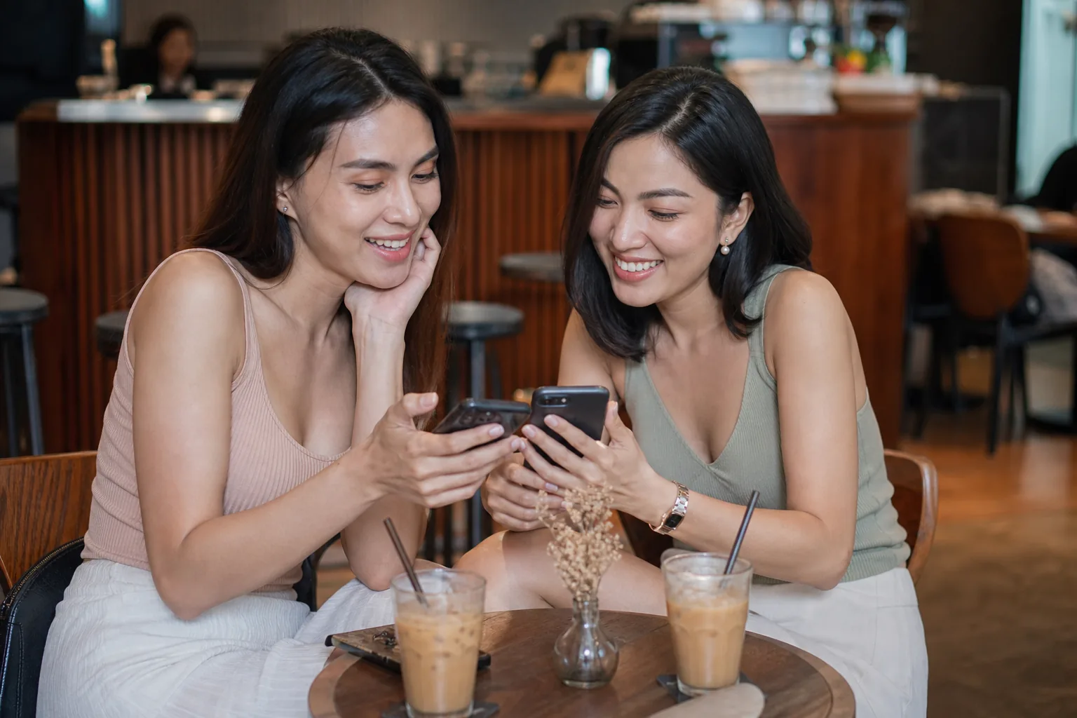 Two Thai trans women friends chatting and reading their phones at a cafe table